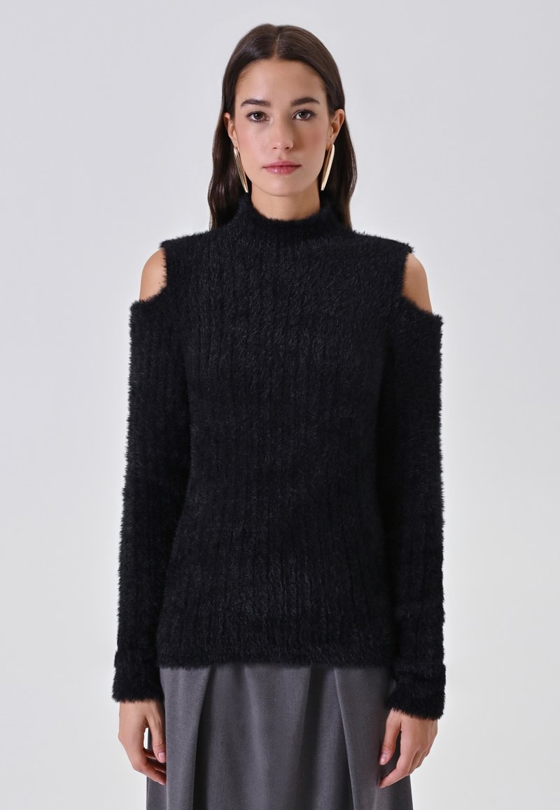 Maglione nero a coste con design a spalla scoperta. Presenta un collo alto e maniche lunghe. Tessuto morbido e texture che conferisce un aspetto accogliente.