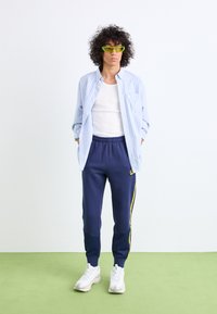Camisa de botão azul e branca com riscas, t-shirt branca por baixo, calças de treino azul-marinho com riscas amarelas nas laterais, e ténis brancos. Óculos de sol verdes como destaque.