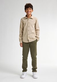 Lichtbeige overhemdsblouse met kraag, gecombineerd met olijfgroene broek en witte sneakers. Soepele textuur, casual pasvorm en zichtbare naden.