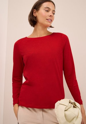 Femme aux cheveux bruns courts, portant un t-shirt rouge à manches longues et un pantalon beige, tenant une veste beige clair dans la main droite.