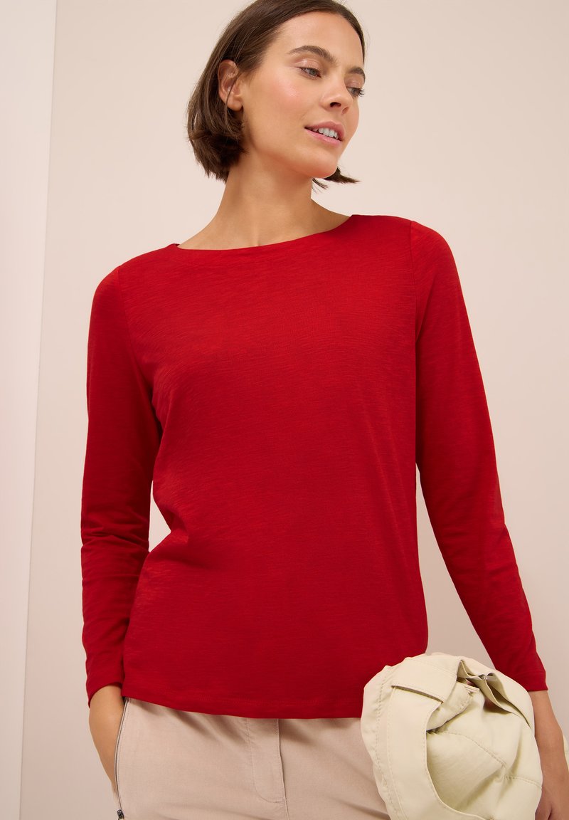 Donna con capelli corti castani, che indossa una maglietta rossa a maniche lunghe e pantaloni beige, tiene in mano destra una giacca beige chiaro.