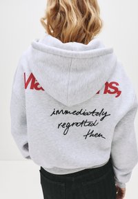 Siva kapuca z gornjim velikim rdečim logom "Vans" na levem ramenu in črnimi ročno napisanimi besedami na hrbtu, izdelana iz mehkega, teksturiranega materiala.