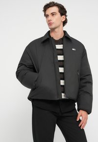 Mustat lyhyet puhvitakki, jossa on vetskari edessä, leveä kaulus ja sivutaskut. Pukeutuneena raidallisen neulepaidan päälle, jossa on sileä, kiiltävä pinta.