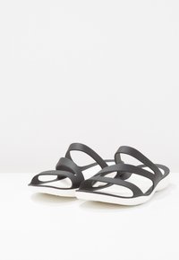 Sandales en caoutchouc noir avec plusieurs brides courbées. Semelle intérieure rembourrée blanche. Semelle légèrement arrondie, présentant des détails découpés dans les brides.