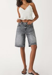Witte, geplooide cropped top met gerilde randen, gecombineerd met hooggetailleerde, knielange zwarte denim shorts met rafelige zoom. Zwarte slippers.
