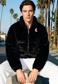 Muškarac tamne kose u crnoj jakni na patentni zatvarač i bijelim hlačama sjedi vani na niskom zidu, u pozadini su palme.