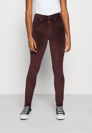 Persona con pantalones de terciopelo de corte ajustado y cintura alta de color púrpura oscuro, top blanco y zapatillas negras y blancas, de pie contra un fondo liso.