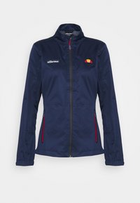 Marinblå vindjacka med hög krage, röda dragkedjedetaljer och en liten logotyp på bröstet. Tillverkad av slitstark, lättviktig tyg.
