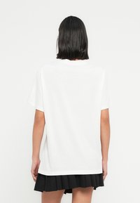 Λευκό βαμβακερό μπλουζάκι με κοντά μανίκια και στρογγυλή λαιμόκοψη, με άνετη εφαρμογή και απλό σχέδιο στο πίσω μέρος. Φορεμένο με μαύρη πλισέ φούστα.