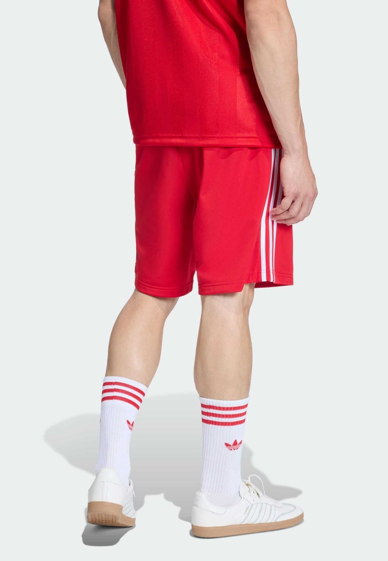 Shorts de sport rouges en tissu léger avec des bandes blanches sur les côtés, assortis de chaussettes blanches à rayures rouges et logo, ainsi que des baskets blanches.