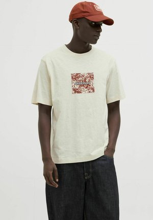 Lys beige T-shirt med et kvadratisk rødt blomstermotiv, der har "Jack & Jones" i midten. Båret med mørke denimbukser.