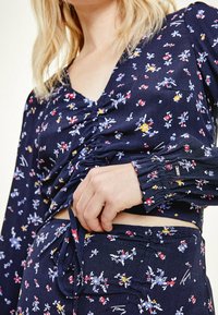 Blusa floreale blu marina con frontale arricciato, maniche lunghe con polsini arricciati e scollo a V. Realizzata in tessuto morbido e leggero.