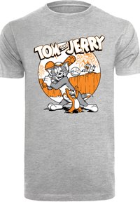Pelēka T-kreklu ar melnu un oranžu zīmēto grafiku, kurā attēloti Tom un Džerijs, ar rotaļīgām detaļām fona žogā un augos.