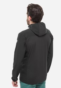 Homme portant une veste à capuche noire et un pantalon turquoise, se tenant de dos contre un fond uni.