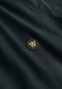 Mustast kangast väike ümmargune pruun plaastriga, mille keskel on õmmeldud põimitud valged tähed "WW".