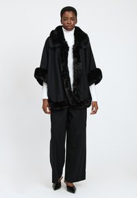 Cape noire avec un ourlet en fausse fourrure peluche sur le col et les poignets, avec un design ample. Portée sur un col roulé blanc et un pantalon noir.