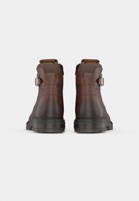 Par de botas de cuero marrón hasta el tobillo con suelas oscuras, hebillas laterales y cuellos acolchados vistas desde atrás sobre un fondo blanco liso.