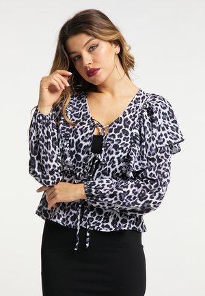 Frau mit langen, welligen Haaren, trägt Creolen und eine schwarz-weiß leopardgemusterte Bluse mit Rüschenärmeln und einen schwarzen Rock.