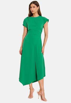 Frau trägt ein grünes asymmetrisches Midikleid mit Kappenärmeln und einem vorderen Schlitz, kombiniert mit nudefarbenen High-Heel-Sandalen.