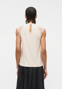 Άτομο με πλεξούδες στα μαλλιά που φοράει αμάνικη εκρού μπλούζα με δαντελένιες λεπτομέρειες στους ώμους και κλείσιμο με κουμπί στο πίσω μέρος.