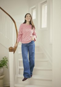 Rosa gestrickter Pullover mit strukturiertem Muster, blaue Weite-Hosen-Jeans und dunkle Schuhe, die auf einer weißen Treppe mit Holzgeländer stehen.
