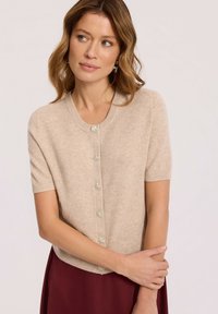 Kobieta w beżowym kardiganie z krótkim rękawem i ozdobnymi guzikami oraz burgundowej spódnicy, patrząca z boku z delikatnie skrzyżowanymi ramionami.