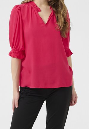 Kvinde iført en lys pink bluse med rynkede V-hals og 3/4 pufærmer, kombineret med sorte bukser, stående med afslappede hænder.