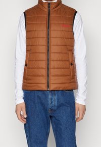 Braune Steppweste mit Stehkragen, Frontreißverschluss und horizontalem Steppmuster. Verfügt über ein rotes Logo und schwarze Seitenakzente.