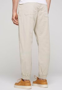 Pantalons de jogging beiges, légers, avec des poignets élastiques, des poches arrière et une coupe décontractée. Portés avec des baskets beiges aux détails lacés.
