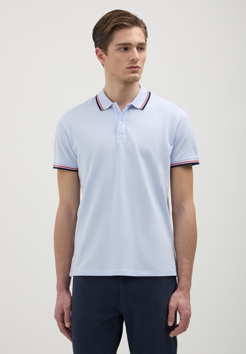 Giovane in camicia polo azzurro chiaro con colletto e bordi delle maniche a righe blu marino e rosso, e pantaloni blu scuro, in piedi contro uno sfondo neutro.