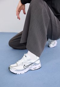 Baskets de sport blanches et argentées avec des matériaux en maille et synthétiques, dotées de lignes d'accent noir, de lacets et de semelles texturées.