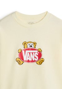 Sudadera de color amarillo claro con un oso de dibujos animados sosteniendo un logo ovalado rojo con la palabra "Vans" en letras blancas. Tejido suave con escote de barco.