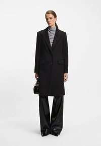 Sort lang frakke med spids lapel og to forlommer, lagdelt over en grå turtleneck, parret med brede sorte plisserede bukser.