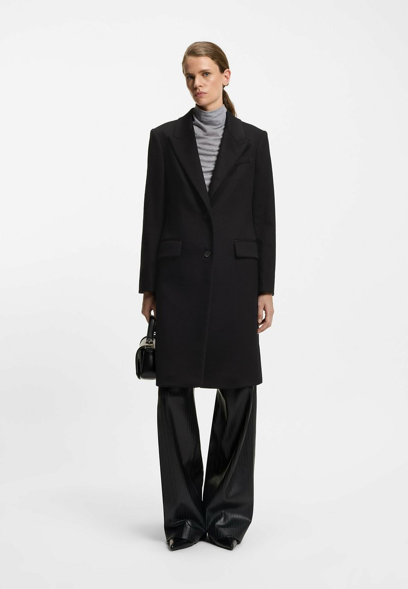 Sort lang frakke med spids lapel og to forlommer, lagdelt over en grå turtleneck, parret med brede sorte plisserede bukser.