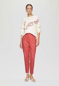 Weißer Strickpullover mit rotem Schriftzug in geschwungener Schrift, kombiniert mit korallenfarbener Slim-Fit-Hose und weißen Riemchensandalen. Glatte Textur und schlichte Silhouette.