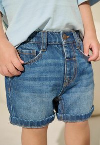 Jeansshorts, hellblau, mit umgeschlagenen Säumen, versehen mit gestickten Details und Taschen. Die Hand des Models ruht auf dem Bund.
