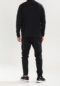 Černá sportovní bunda s vysokým límcem, doplněná o ladící zúžené kalhoty. Materiál působí hladce; má boční kapsy. Černé sportovní boty.