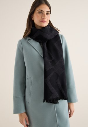 Woman wearing a light blue coat and long black scarf with geometric patterns, standing against a plain light background.