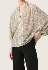 Blusa leggera color verde tenue con motivo, scollo a V, maniche ampie e vestibilità rilassata, indossata sopra pantaloni neri. Texture sottile visibile.