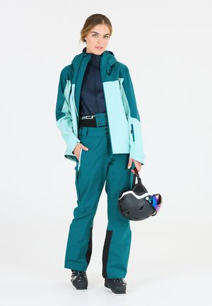 Teal og lyseblå skijakke med lynlås-lommer, parret med matchende teal bukser. Modellen holder en sort hjelm med spejlvisir.