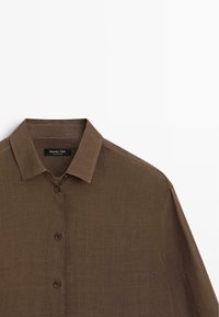 Braunes langärmliges Hemd mit Knopfleiste und klassischem Kragen, aus strukturiertem Stoff, vor weißem Hintergrund präsentiert.