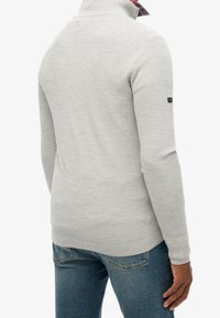 Pull en maille gris clair avec un col montant et une finition texturée, arborant un col intérieur à carreaux. Coupe décontractée, associé à un jean en denim bleu.