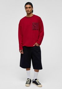 Rode longsleeve shirt met een gestreepte borstzak, gecombineerd met zwarte shorts, witte sokken en zwarte sneakers. Eenvoudig, casual ontwerp.