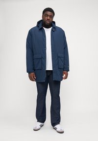 Parka blu navy con cappuccio e chiusure a pressione, due tasche frontali e una vestibilità rilassata. Indossata sopra una camicia bianca e abbinata a jeans scuri e sneakers.