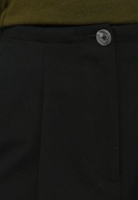 Pantalons en tissu noir avec une texture lisse et un léger côtelé diagonal, dotés d'un bouton noir avec logo gravé et de poches latérales.