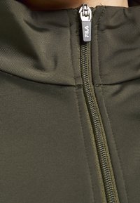 Oliwkowa kurtka na zamek błyskawiczny o fakturowanej tkaninie, z metalowym zamkiem i metką z logo. Zbliżenie na kołnierz i strefę zamka.