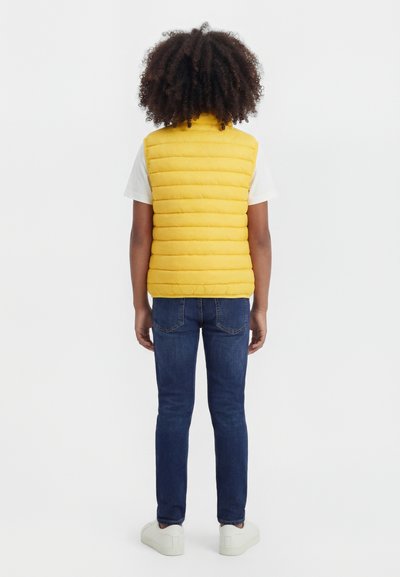 Child with curly hair wearing a yellow puffer vest, white t-shirt, blue jeans, and white sneakers, standing facing away on a white background.