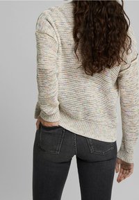 Leicht strukturierter beiger Pullover mit mehrfarbigen Sprenkeln, gerippte Bündchen und Saum, kombiniert mit dunkelgrauen, eng anliegenden Jeans. Von hinten betrachtet.