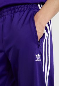 Violet sporta bikses ar baltām sānu svītrām. Iezīmes ietver elastīgu jostasvietu un izšūtu Adidas logo kreisajā apakšā.