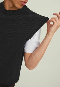Schwarzes gestricktes ärmelloses Oberteil mit hohem Rippkragen, aus strukturiertem Stoff und lockerer Passform, über einem weißen Hemd getragen.